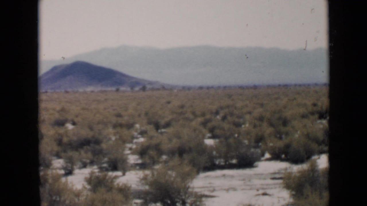 1949 San Diego-area 8mm frame showing sandy scrubland, a low hill, and layered mountains under a pale sky.