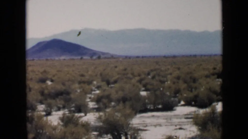 A steady 8mm view of San Diego desert scrub with a dark rounded hill and hazy mountains in the background.