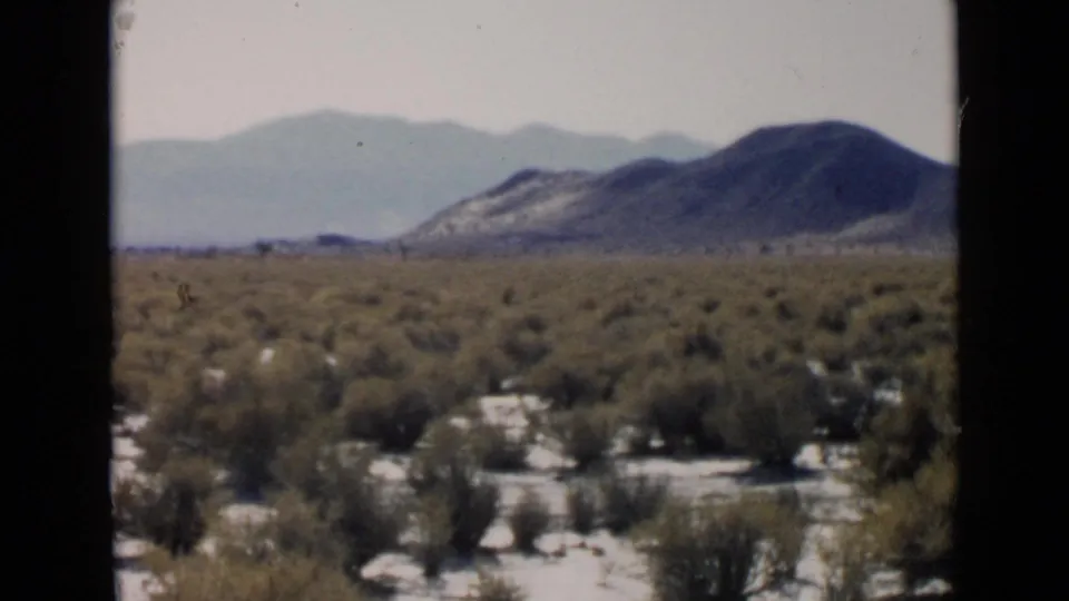 1949 home-movie frame of open San Diego desert terrain with scattered shrubs, a broad hill, and muted mountains beyond.
