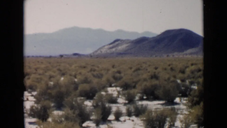 San Diego-area 1949 8mm frame showing shrubland, pale sand, a broad hill, and layered mountains under haze.