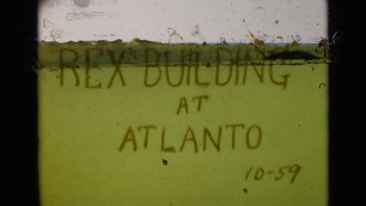 8mm frame of a handwritten green title card reading Rex Building at Atlanto, with a small 10-59 date and visible film wear.