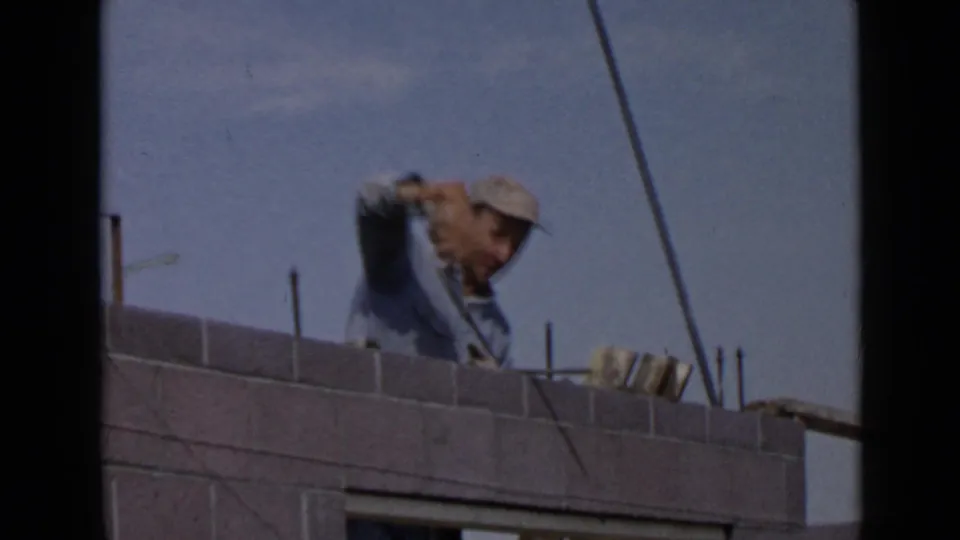 In the San Diego home-movie clip, a capped construction worker appears to apply mortar on a concrete block wall beneath a clear blue sky.