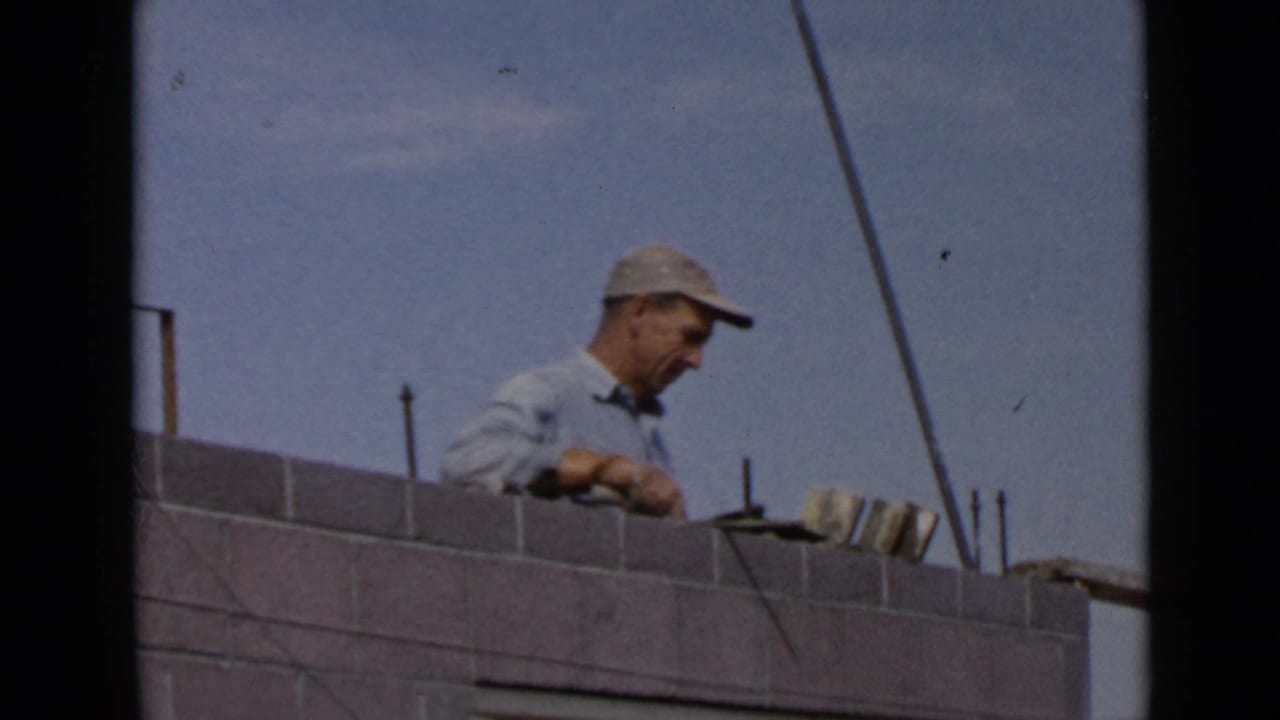 1949 San Diego home-movie frame showing a capped laborer applying cement along the top of a grey brick wall.