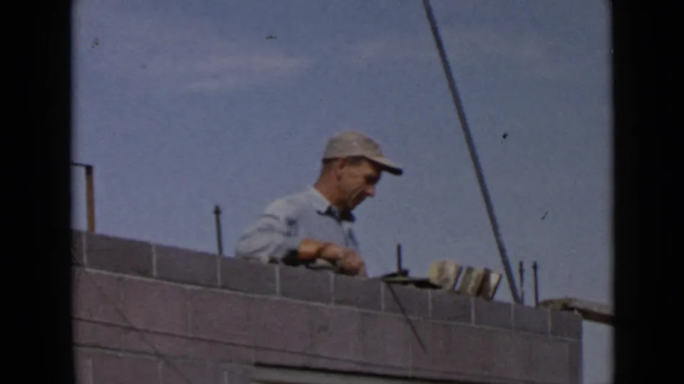 1949 San Diego home-movie frame showing a capped laborer applying cement along the top of a grey brick wall.