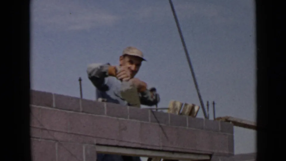 1949 San Diego home-movie still of a laborer leaning over a grey brick wall during masonry work.