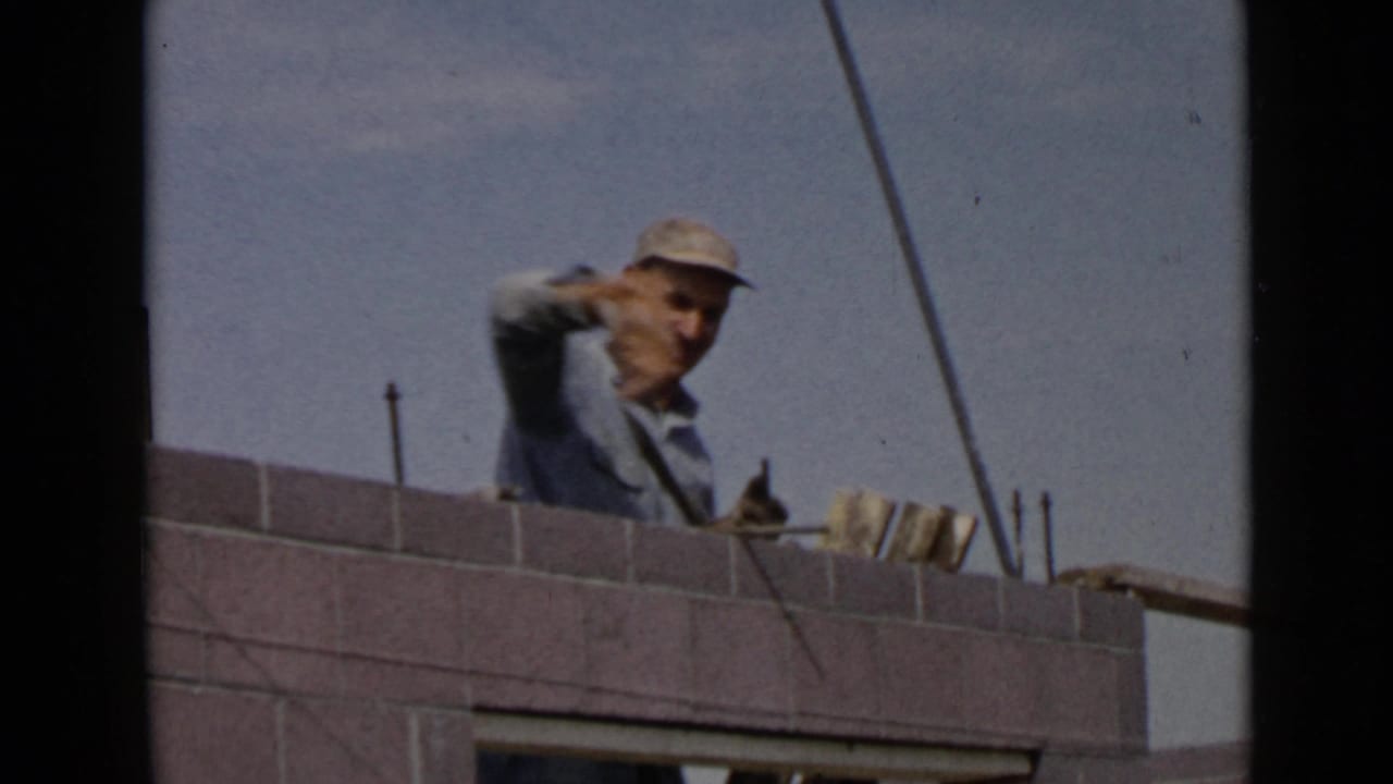 1949 San Diego home-movie frame showing a mason behind a concrete block wall with a trowel and rebar visible under open sky.