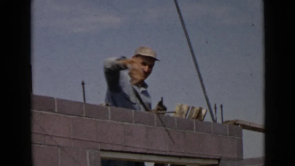 1949 San Diego home-movie frame showing a mason behind a concrete block wall with a trowel and rebar visible under open sky.