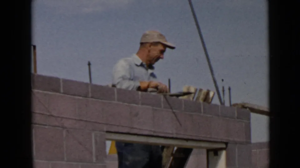 1949 San Diego home-movie frame of a mason focused on trowel work along a partially built concrete block wall.