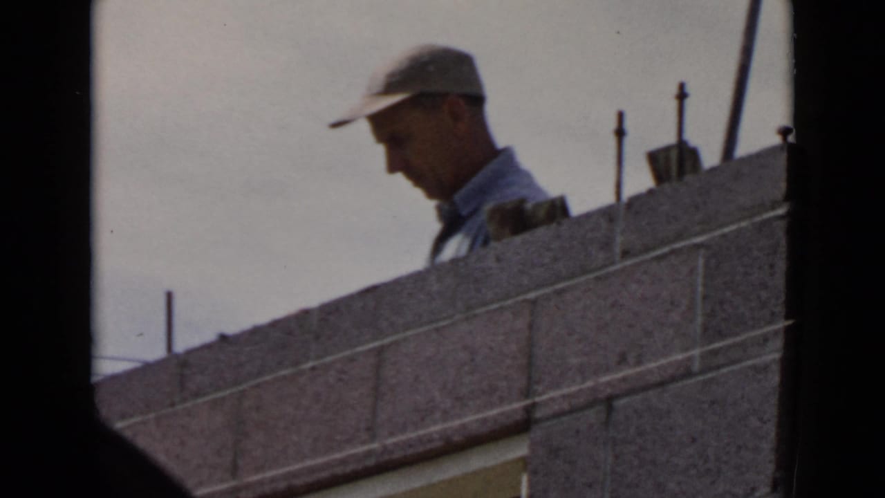 1949 San Diego frame of a capped worker looking down while standing behind a concrete block wall.