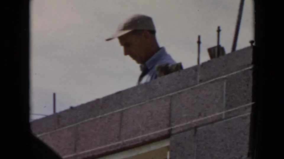 1949 San Diego frame of a capped worker looking down while standing behind a concrete block wall.