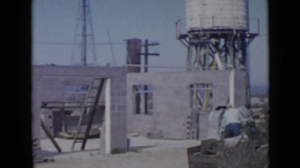 1949 San Diego home-movie frame showing a cinder block construction site with a ladder, raised water tank, and adults seen from behind.