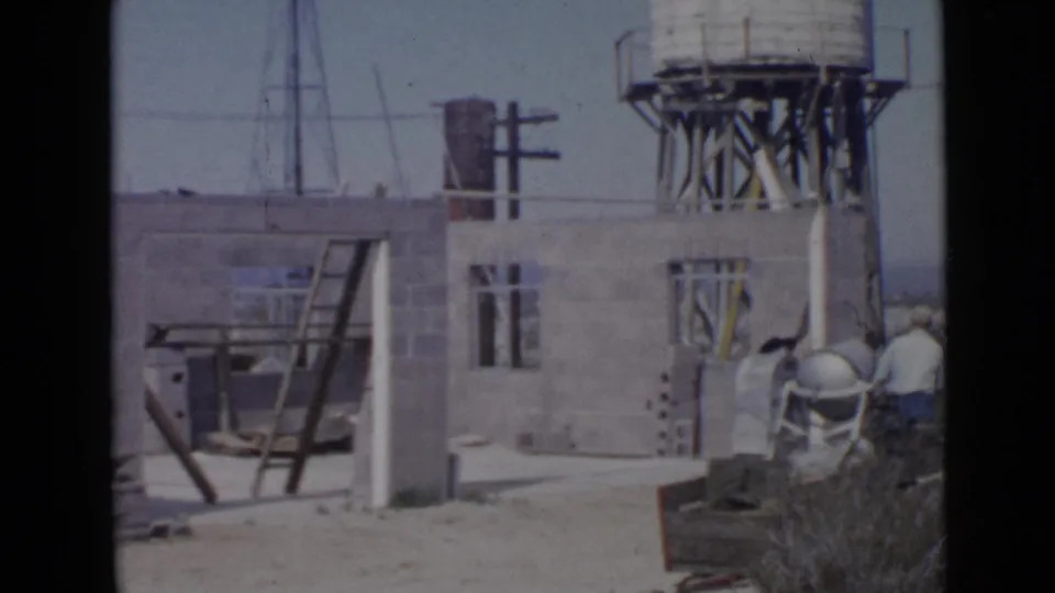 Cinder block construction scene in 1949 San Diego with a raised water tank, ladder, open wall sections, and adults working at frame right.