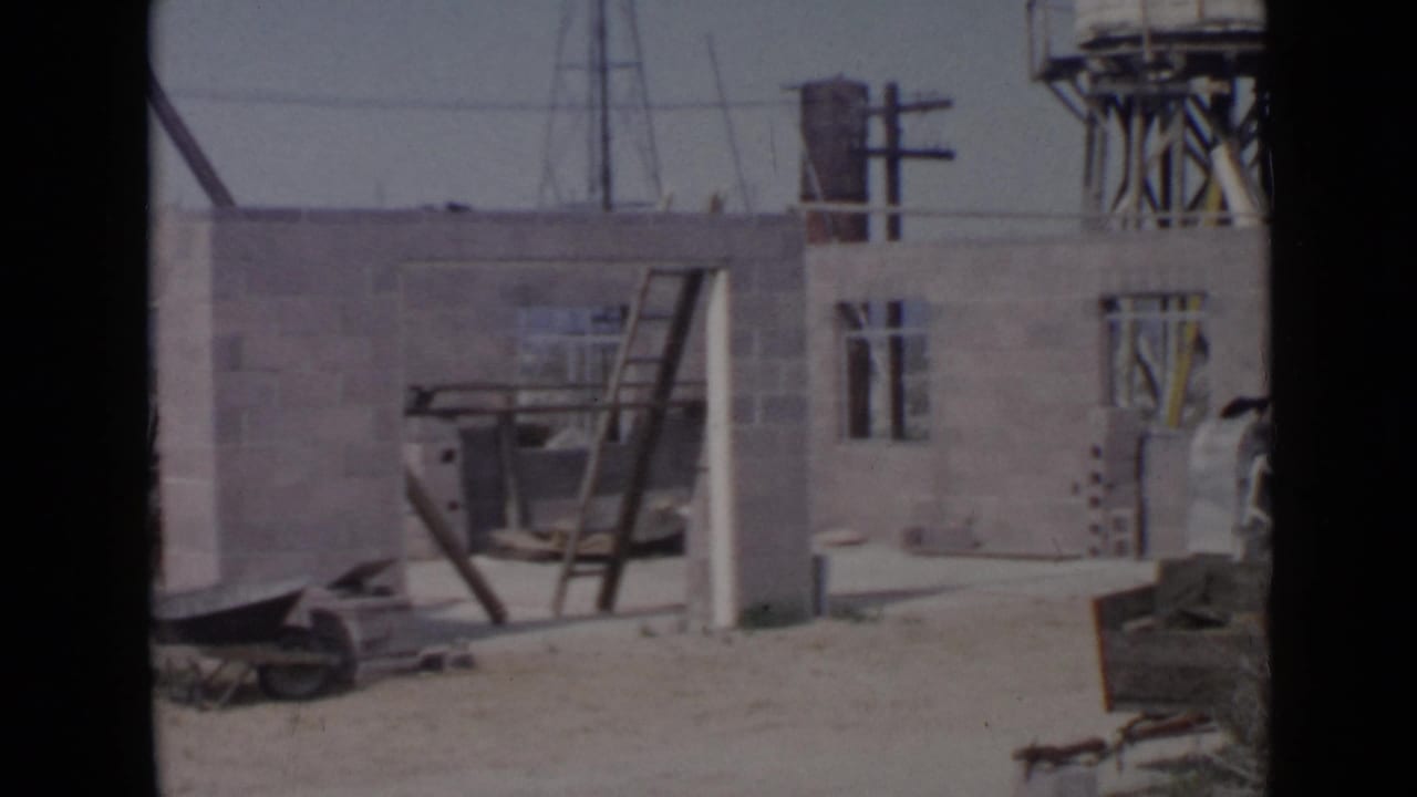 Unfinished block building with ladder, wheelbarrow, utility poles, and water tower in the background.