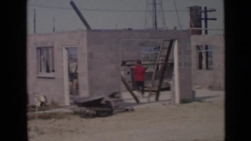 A worker at a 1949 San Diego construction site stands beside a ladder, with block walls, window openings, and poles behind.