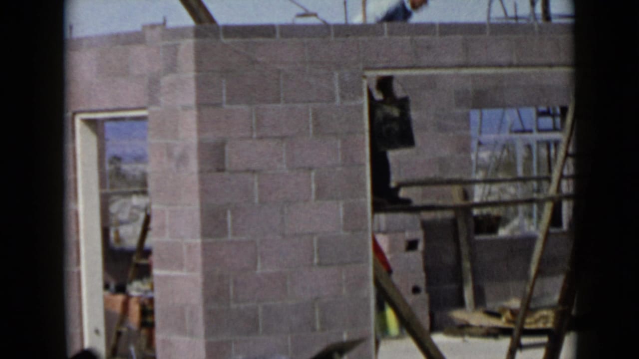 1949 San Diego home-movie frame showing a construction worker partly above a cinder block wall with ladder and framing nearby.