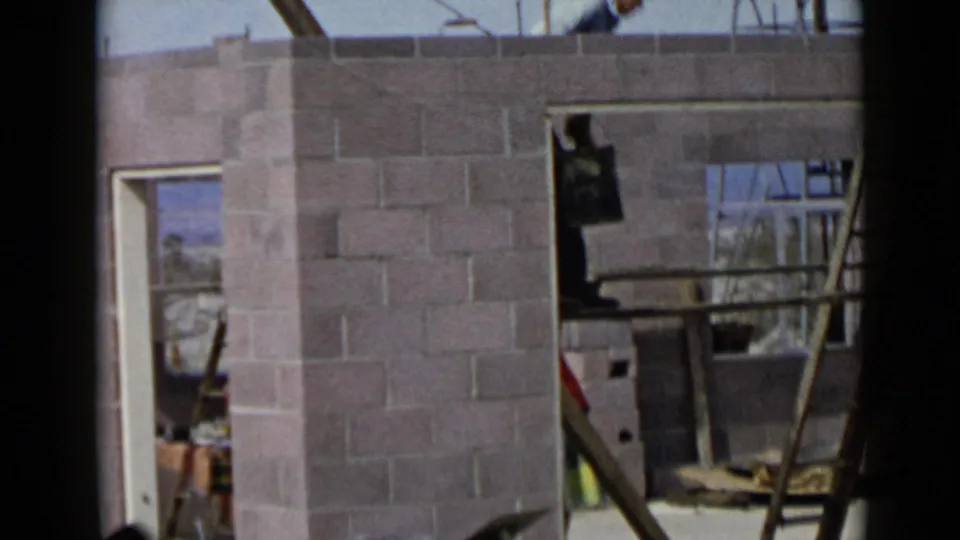 1949 San Diego home-movie frame showing a construction worker partly above a cinder block wall with ladder and framing nearby.