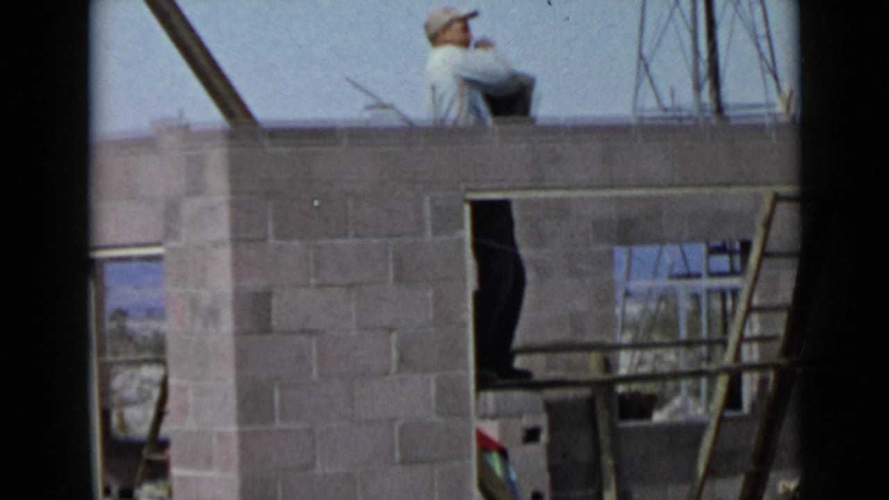 1949 San Diego frame with a capped worker visible above a cinder block wall, surrounded by ladder rails and unfinished framing.