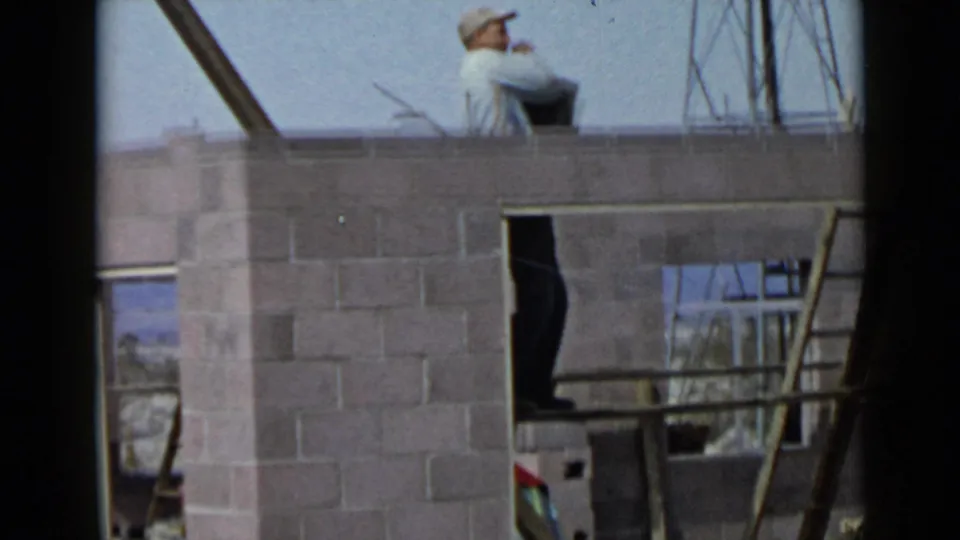 1949 San Diego frame with a capped worker visible above a cinder block wall, surrounded by ladder rails and unfinished framing.