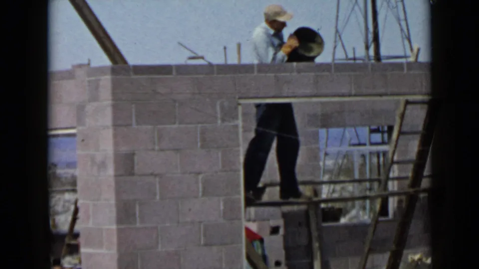 1949 San Diego construction scene showing a worker on a ladder holding a dark bucket beside a partially built cinder block wall.