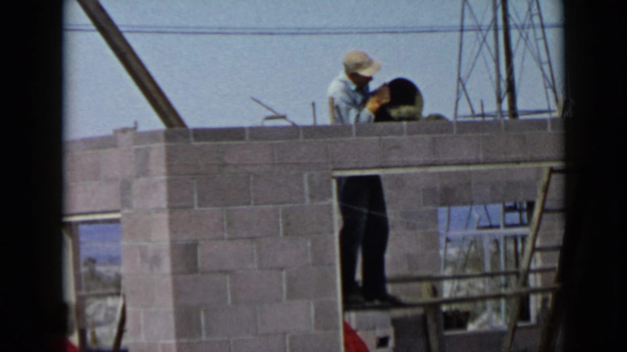 In 1949 San Diego footage, a capped worker lays mortar on a cinder block wall with utility lines and framing behind him.