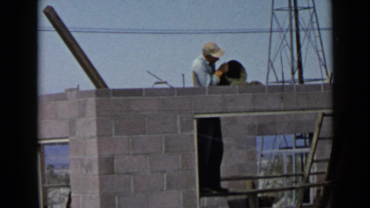A 1949 San Diego home-movie frame shows the worker leaning over a block wall while a dark figure or shadow appears in an opening below.