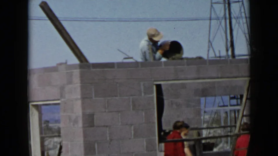 The San Diego construction scene shows a worker applying mortar atop the cinder block wall as another person in red is briefly visible below.