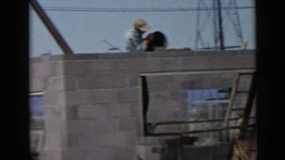 A soft 8mm frame shows the worker bent over the top course of the wall, with a window opening, rails, and background utility lines visible.
