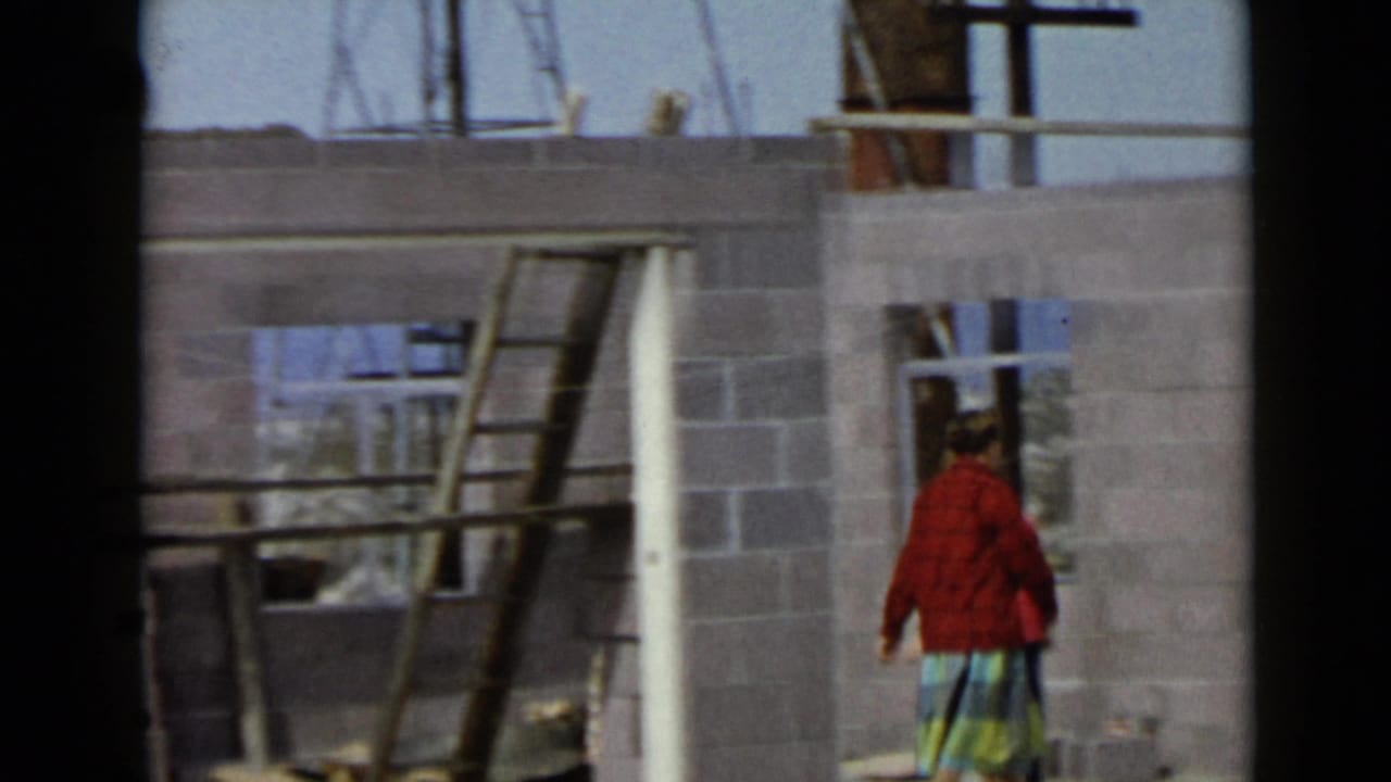 In 1949 San Diego home-movie footage, a woman in a red sweater and plaid skirt stands near open framing and cinder block walls.