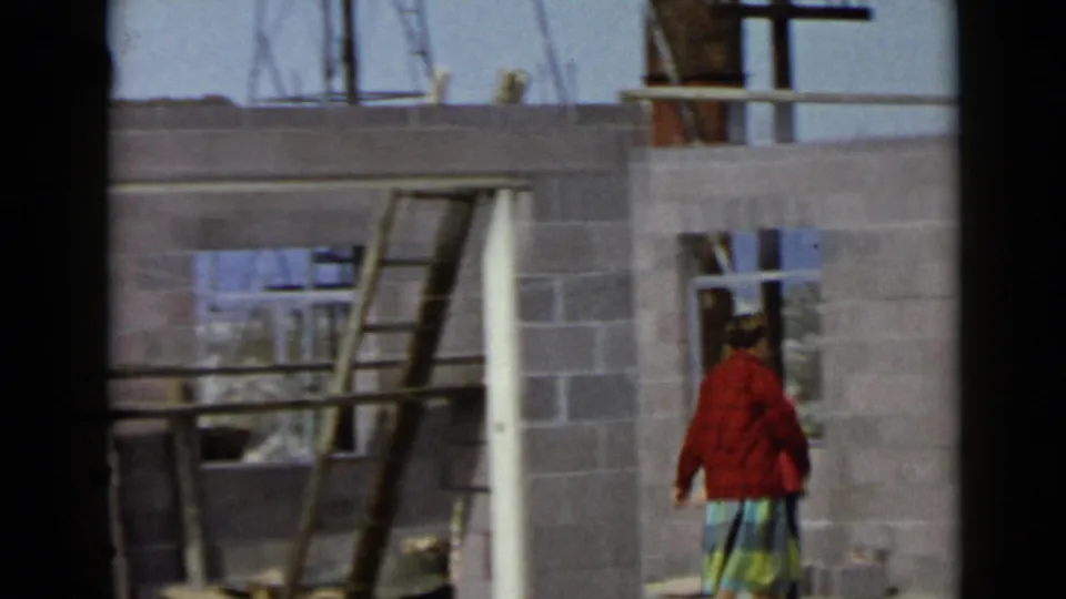 In 1949 San Diego home-movie footage, a woman in a red sweater and plaid skirt stands near open framing and cinder block walls.