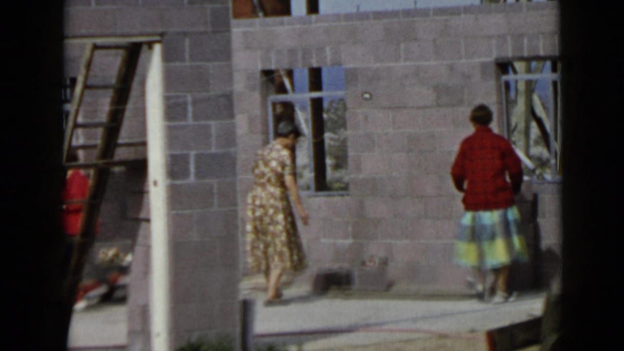 Two women appear at a 1949 San Diego construction site, one in a floral dress and one in a red sweater near unfinished walls.