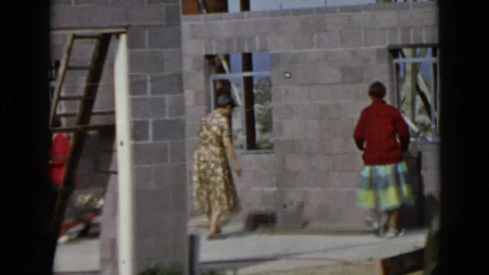 Two women appear at a 1949 San Diego construction site, one in a floral dress and one in a red sweater near unfinished walls.