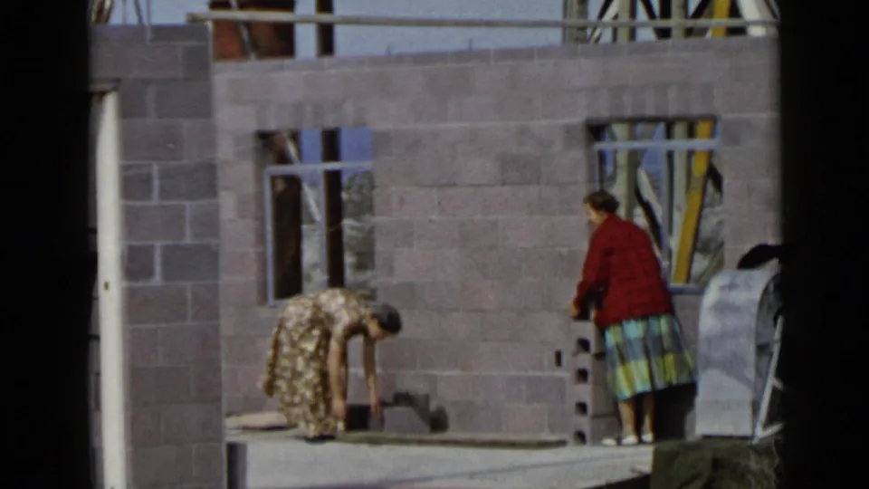 At the San Diego construction site, a woman in a floral dress bends toward a wooden board as another woman watches nearby.