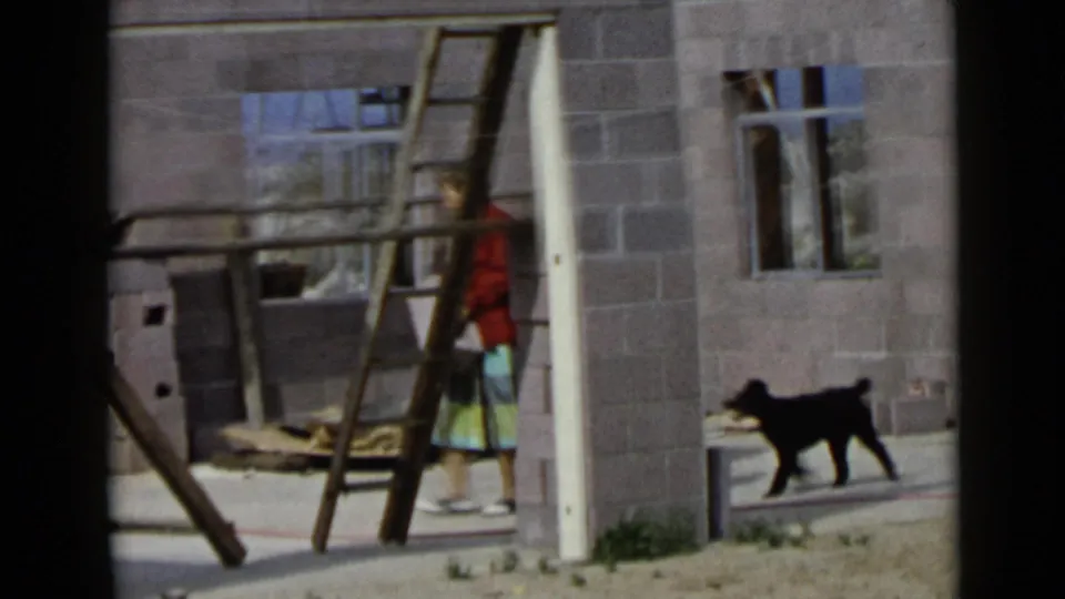 1949 San Diego home-movie frame showing a woman in a red jacket and skirt with a black dog inside an unfinished block structure.