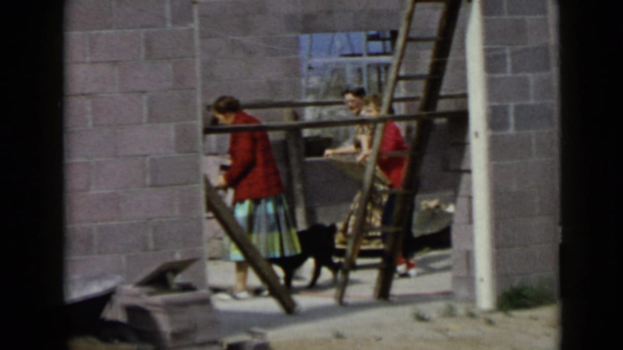 1949 San Diego frame with two women near wood ladders and a black dog in an unfinished cinder block building.