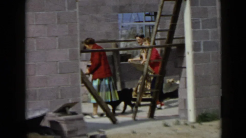 1949 San Diego frame with two women near wood ladders and a black dog in an unfinished cinder block building.