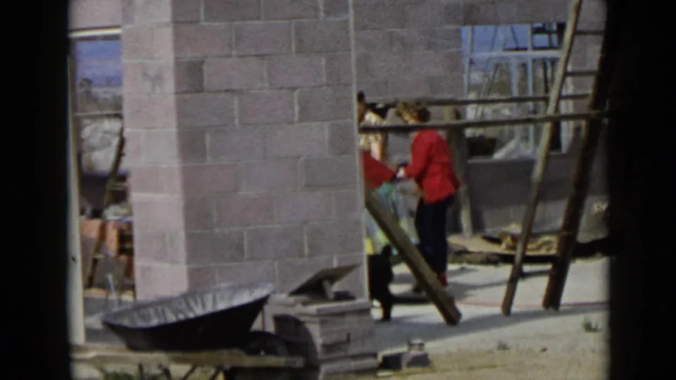 1949 San Diego home-movie frame showing women partly hidden by unfinished block walls, with ladders and a wheelbarrow nearby.