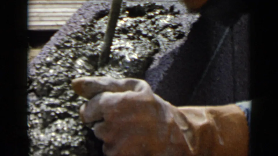 Close 1949 home-movie detail of a hand using a tool on wet cement, with coarse texture and shallow focus.