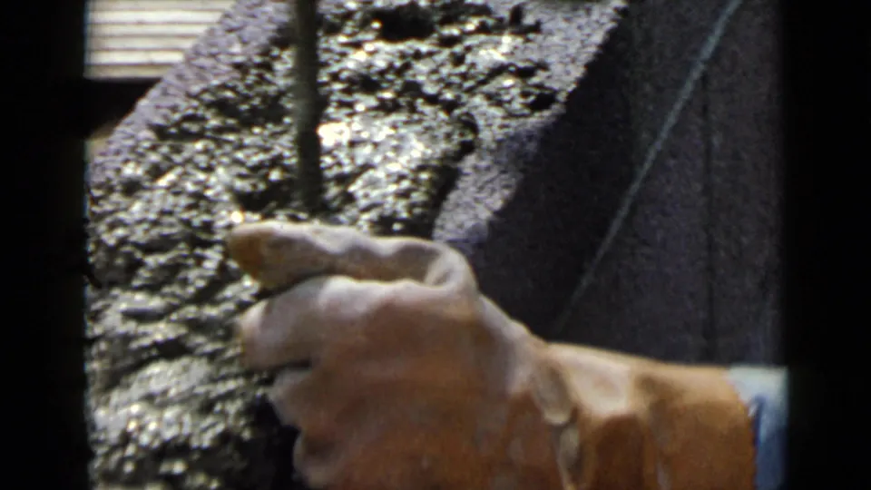 1949 San Diego home-movie frame showing a worker's hand guiding a trowel near wet cement on a rough concrete block.