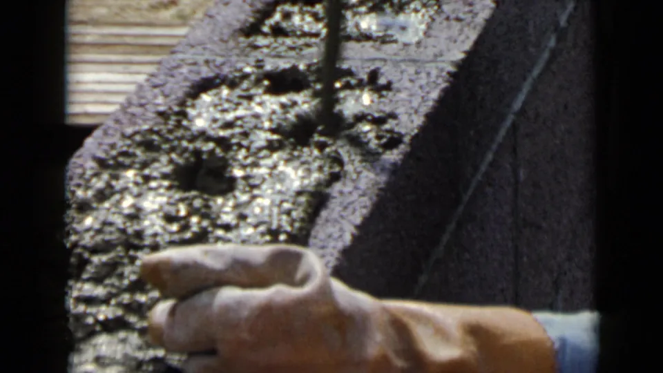 Home-movie frame from the 1949 San Diego clip showing a hand near a trowel and freshly applied cement on a block.