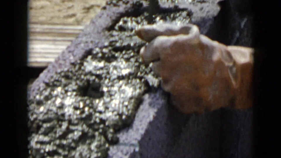 1949 San Diego home-movie frame showing a hand using a small tool to spread wet cement across a cinder block surface.