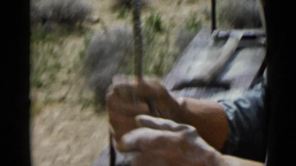 A worker's hands hold a narrow tool outdoors in the San Diego 1949 home-movie footage, with dry brush and work materials behind.