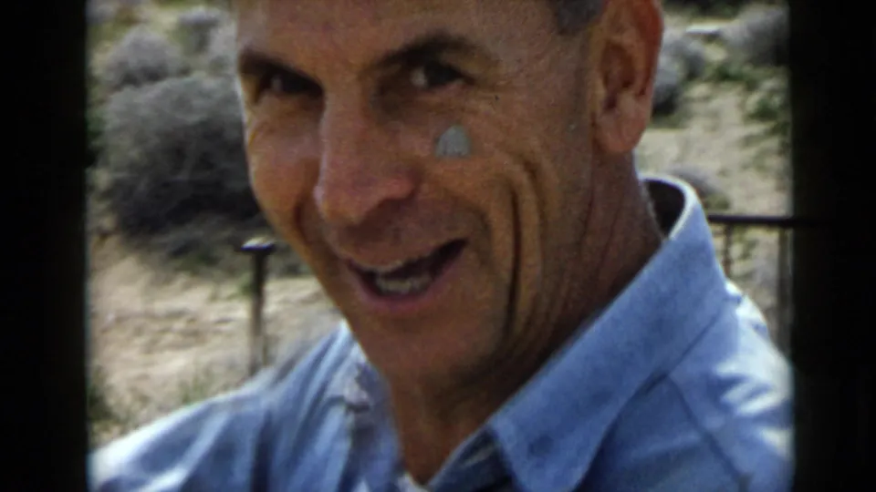 The adult worker smiles toward the camera in a 1949 San Diego home-movie frame, with outdoor brush and work materials behind him.