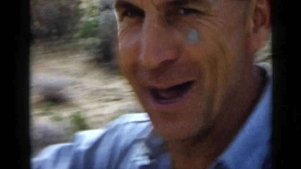 1949 San Diego home-movie frame showing the man smiling outdoors, with sandy soil and sparse vegetation in the background.