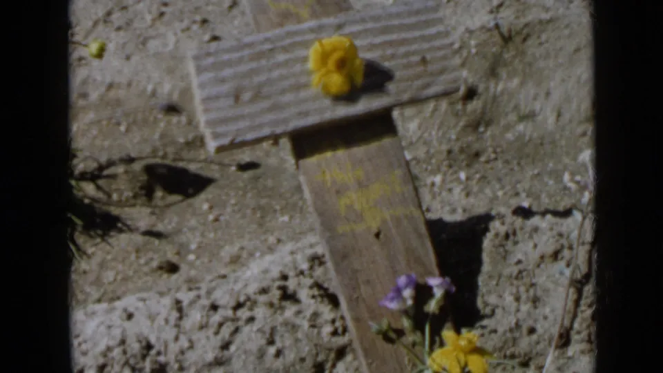 1949 San Diego-area 8mm frame showing a small wooden cross decorated with yellow and purple flowers in dry sandy soil.