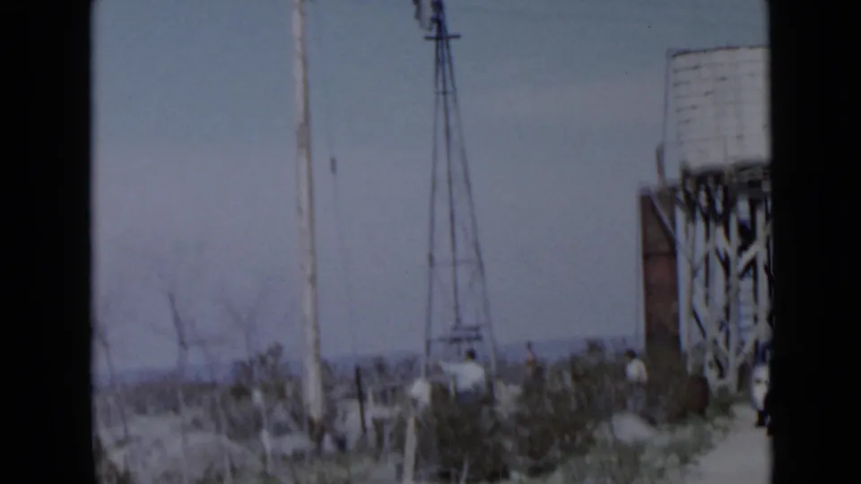 1949 San Diego-area rural scene with a tall metal windmill, elevated water tank, scrub vegetation, fence posts, and distant people.