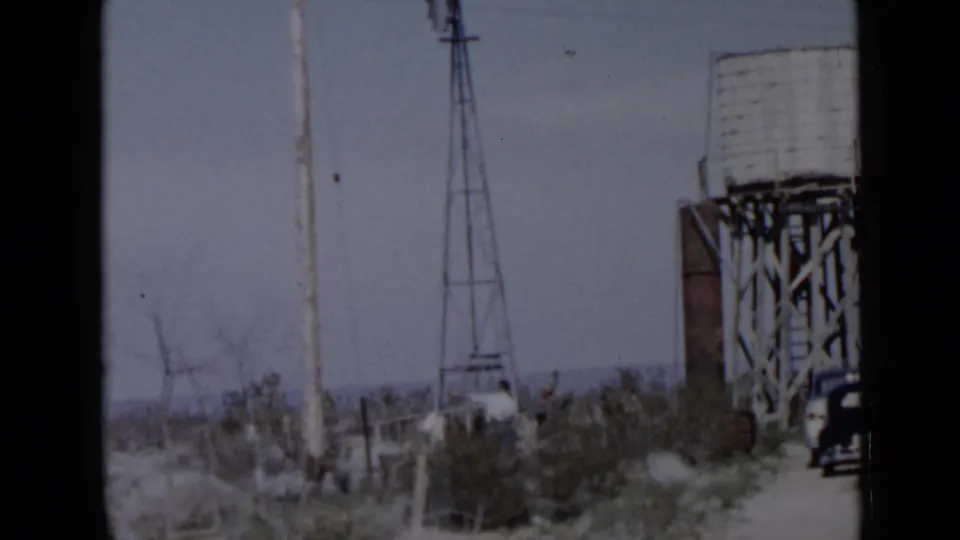 Rural 8mm frame showing a windmill, elevated water tank on wooden supports, sparse vegetation, and people near the structures.