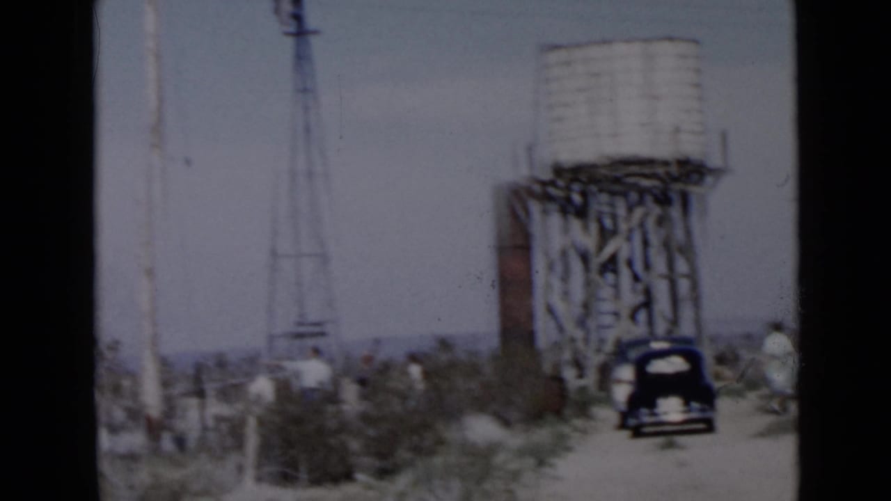 1949 San Diego-area home-movie frame showing a wooden water tower, black car, scrub desert, and small distant people near a dirt road.