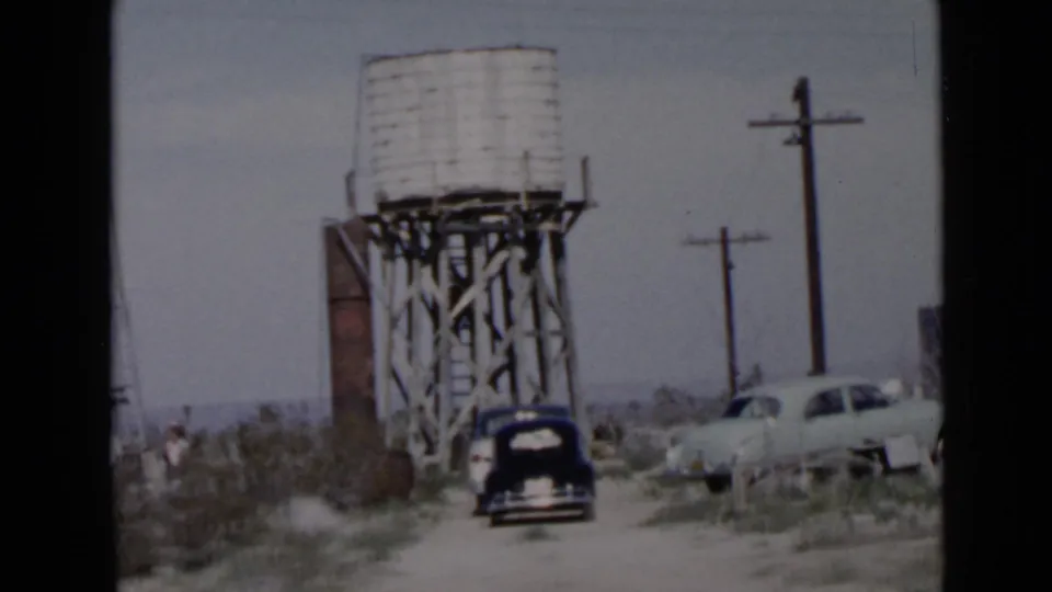 1949 San Diego-area desert campsite view with a wooden water tower, parked black car, light sedan, utility poles, and dry brush.
