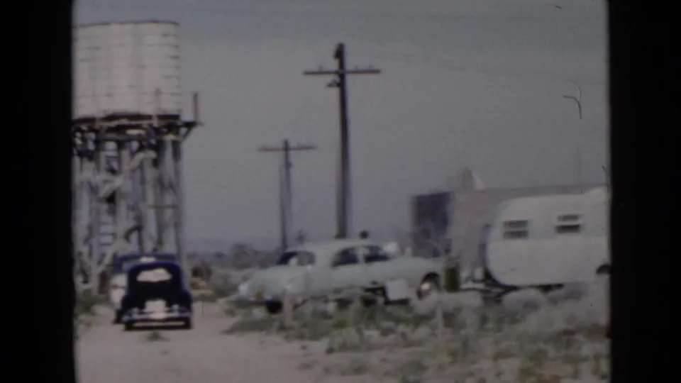 1949 San Diego-area frame showing a dirt road campsite with black car, light sedan, white travel trailer, utility poles, and desert brush.