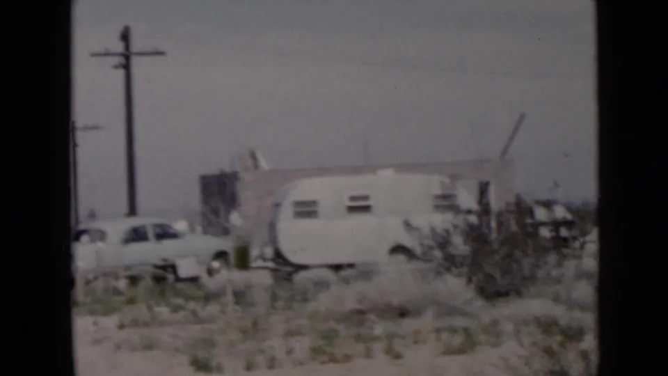 1949 San Diego-area home-movie frame focused on a white travel trailer, light sedan, utility poles, and scrub desert surroundings.